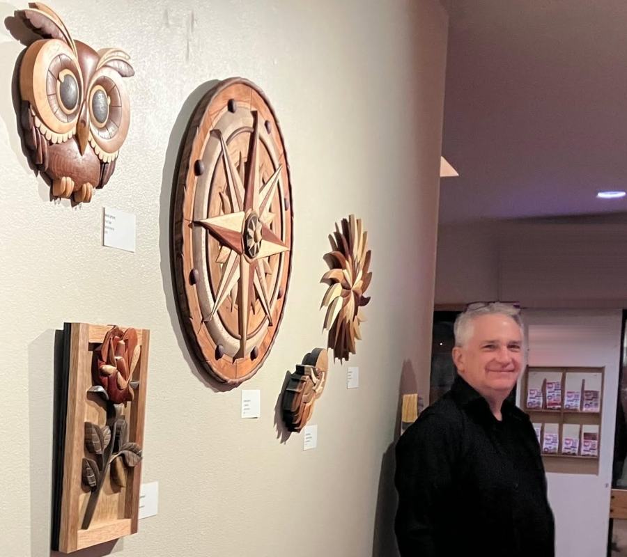Mark Gifford standing beside a wall of his intarsia art, including an owl, compass rose and rose